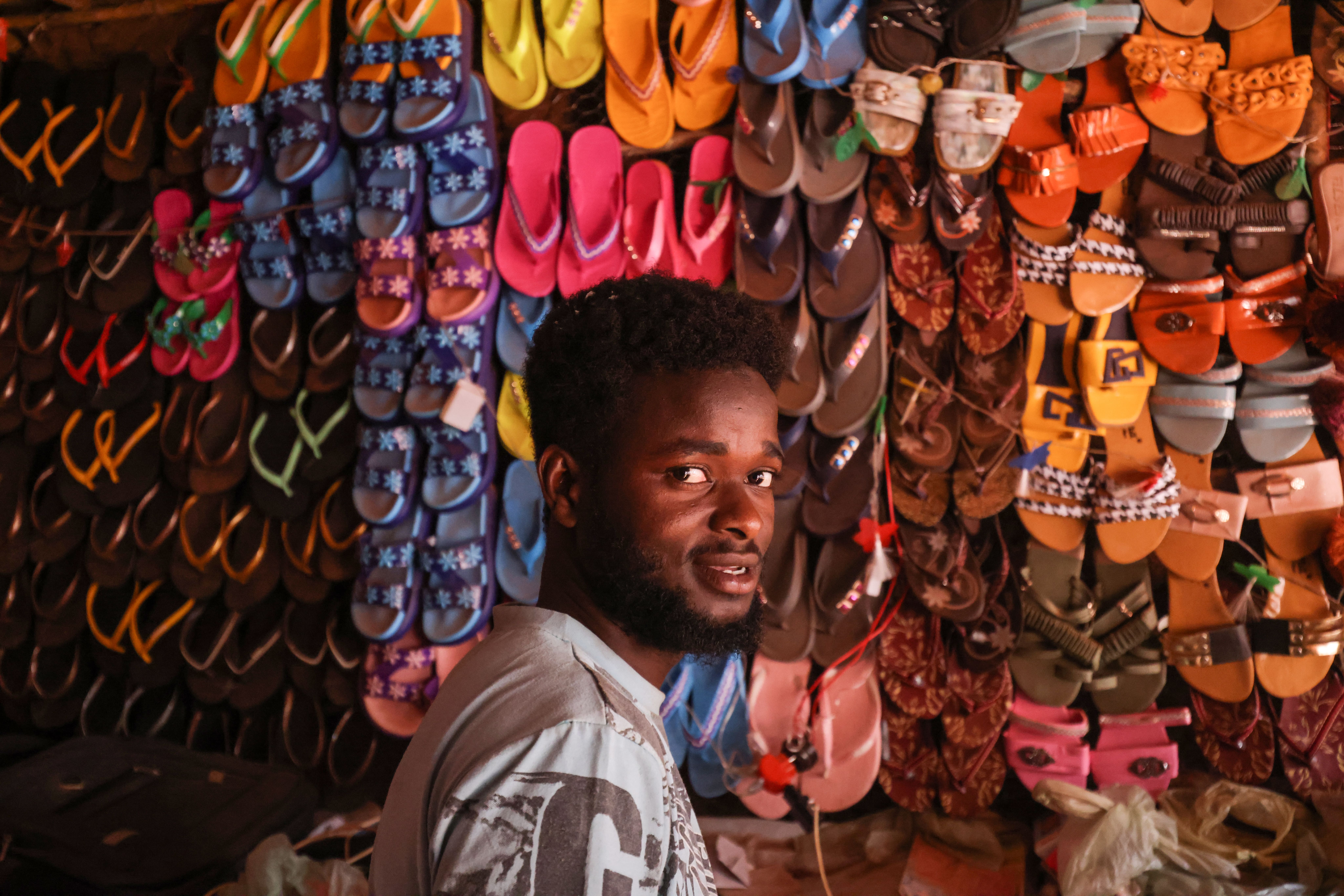 Mussa está na sua loja, em frente a uma parede de filas de sandálias coloridas.